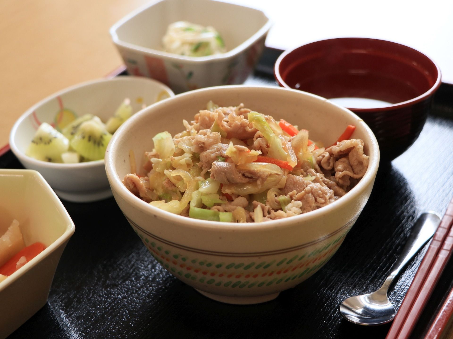 食事の一例である。黒いプレートに丼ものと汁物、小鉢物とデザートが添えられ、お箸とスプーンとともに並べられている。