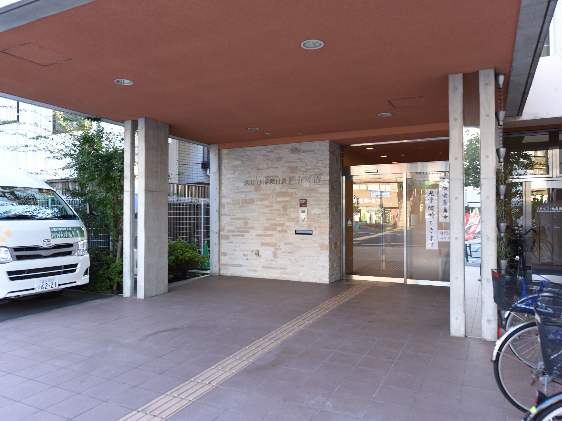 広くかつ明るい色調の正面玄関口。白いバンが停車している。自動ドアの左横には施設名がある。見学が随時可能であるという掲示がある。