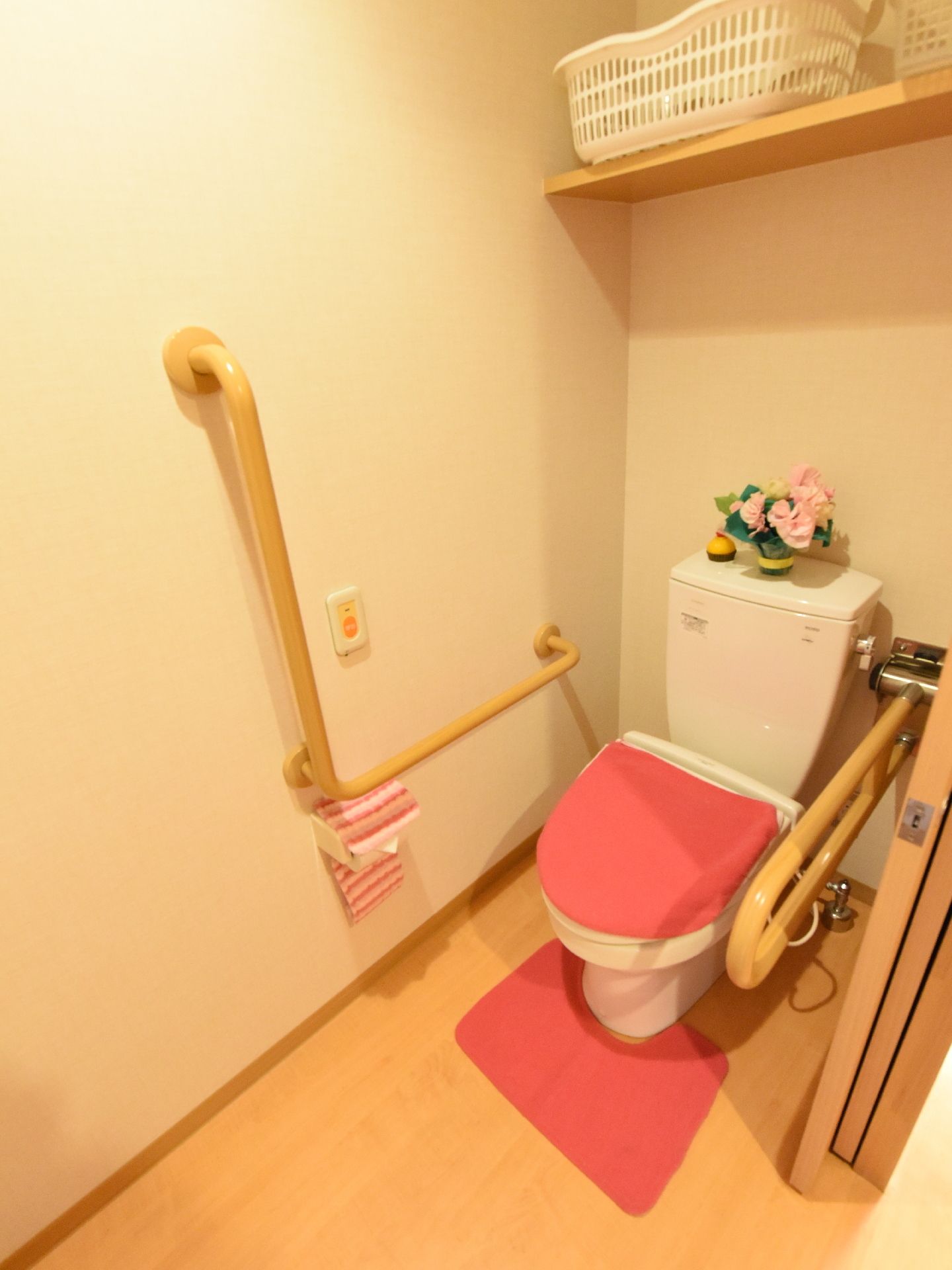 手すりが設置されたトイレスペース。ピンク色の花で装飾がされている。便器、ペーパーホルダー、マットいずれもピンク系統の色で統一されている。