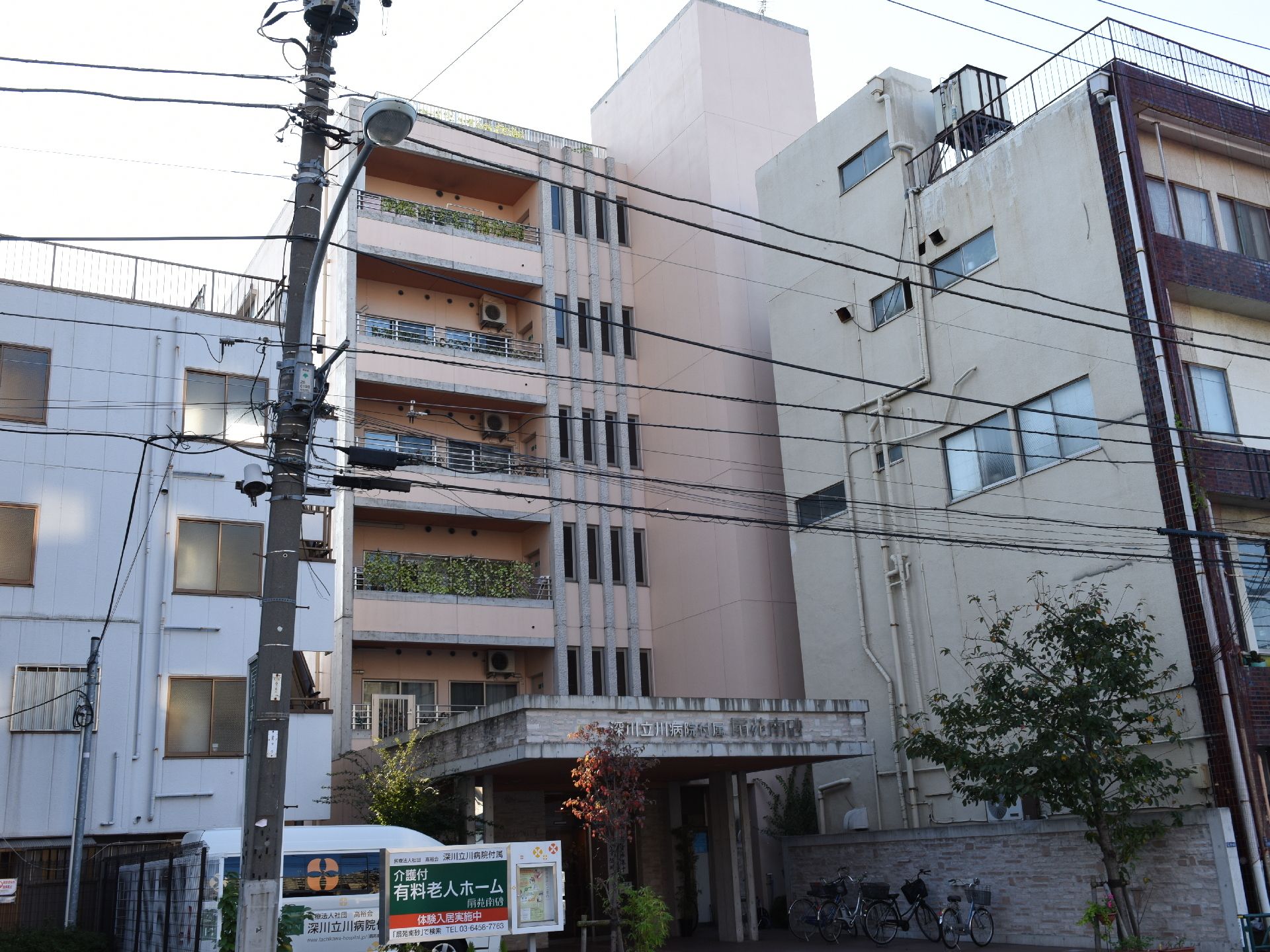 玄関口には施設名が書いてある。電柱と電線、街灯がある。玄関口のある建物は6階建てで、居室にはそれぞれベランダがある。