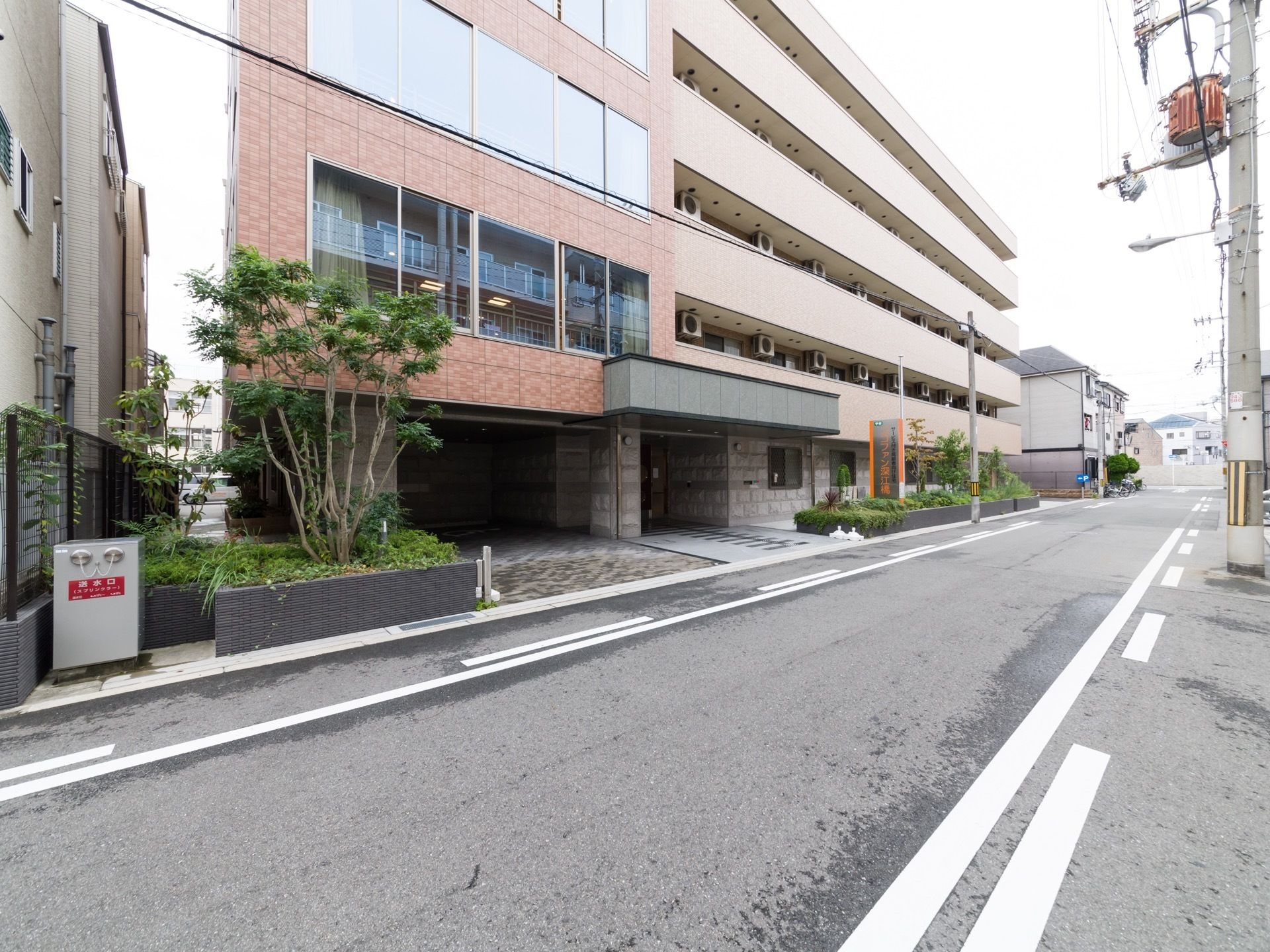 真っ直ぐに伸びた道路沿に位置する建物である。玄関向かって右側には、ベランダのフェンスが突き出ている。建物前方の端に送水口を設置している。