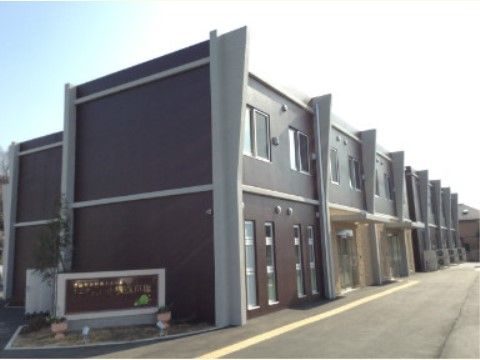 日当たりの良いグループホームである。建物奥の外壁沿いに、エアコンの室外機が複数置かれている。施設前はアスファルトで舗装されている。