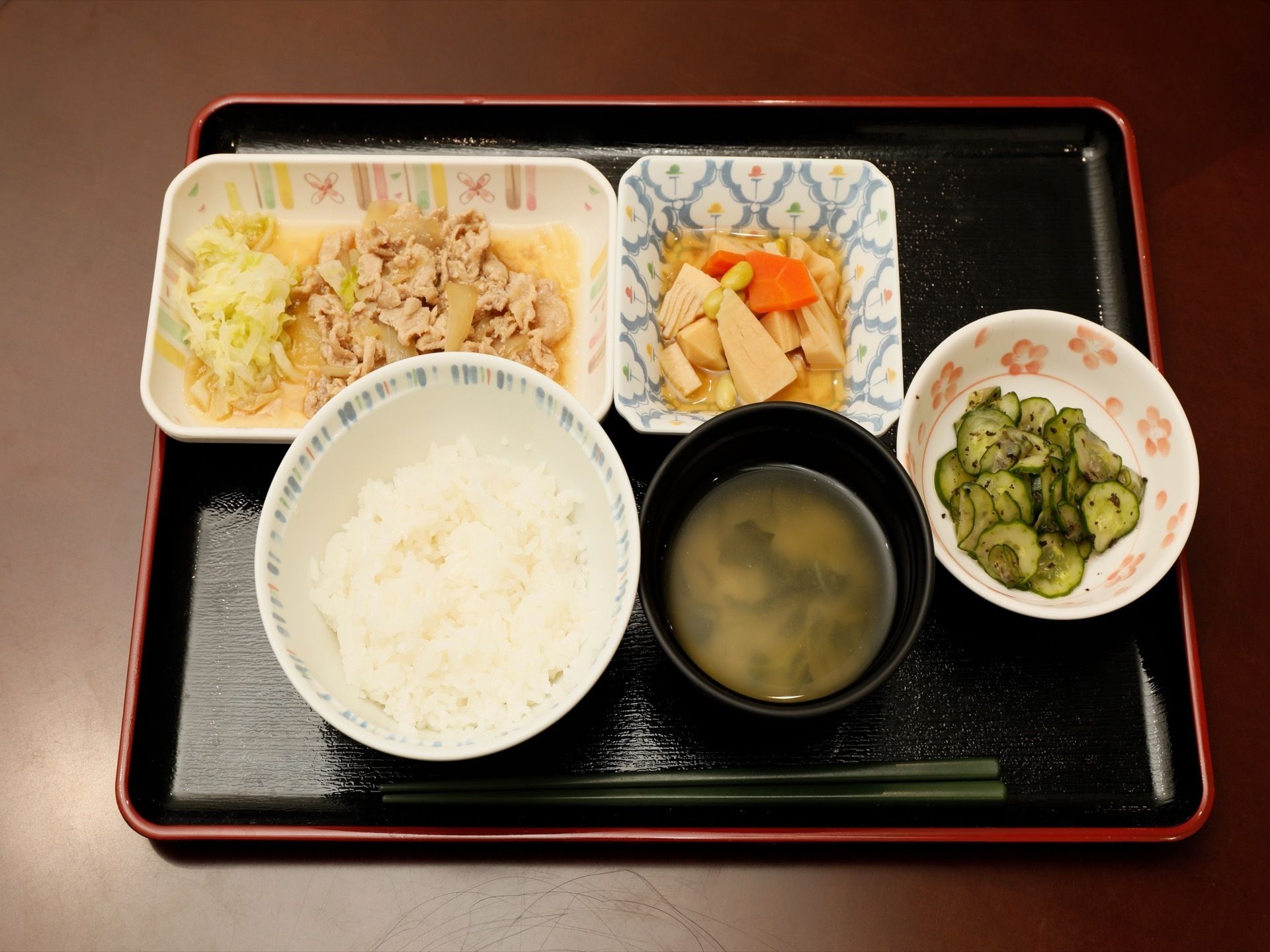 この日の食事がお皿に盛りつけられお盆に並べられている。肉の炒め煮や根菜の煮物、酢の物などが用意されている。