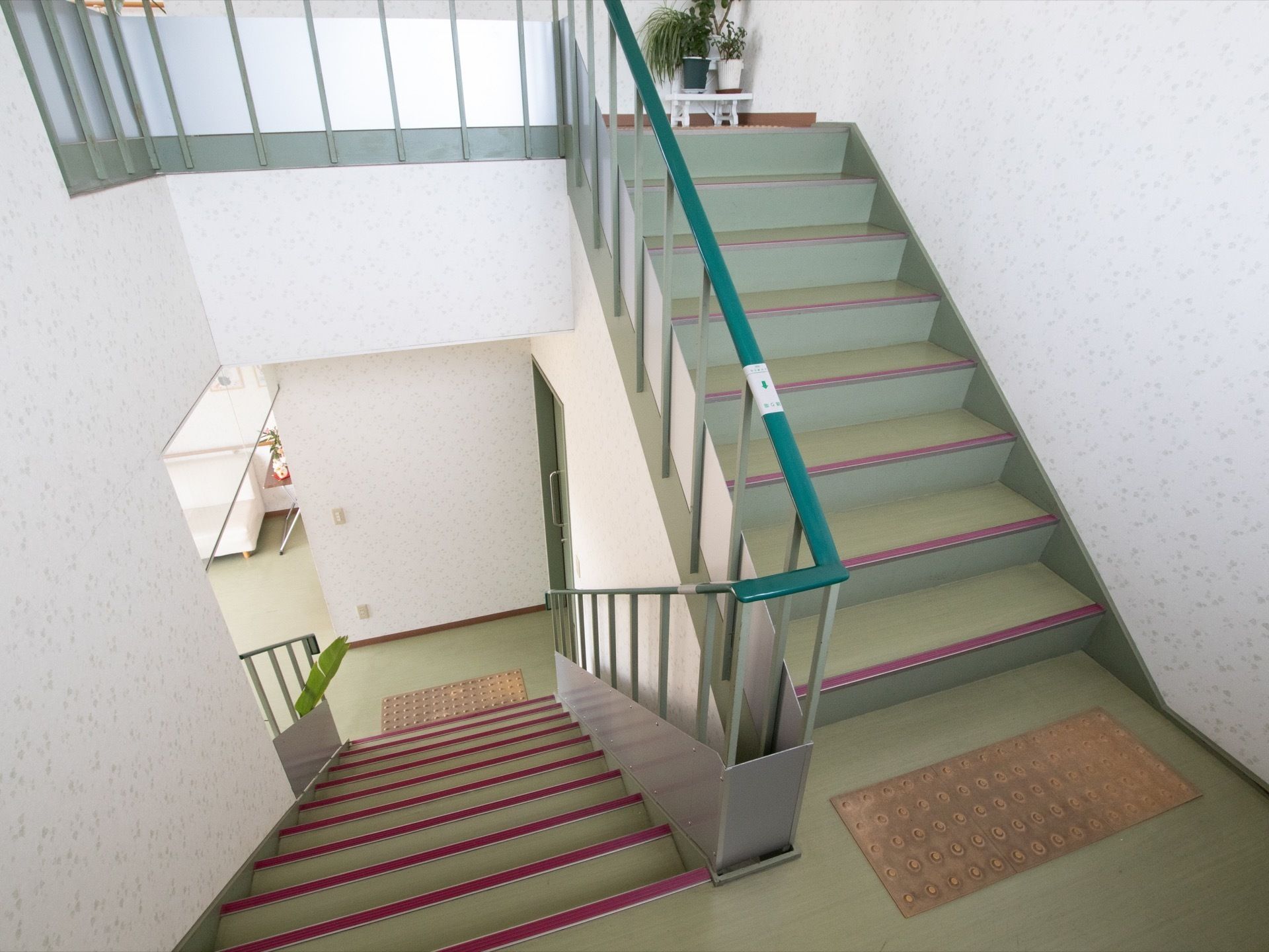 階段には手すりが付き、周りの壁は白い。各段の淵にはエンジ色の滑り止めが付いており、踊り場に観葉植物の鉢植えが飾られている。