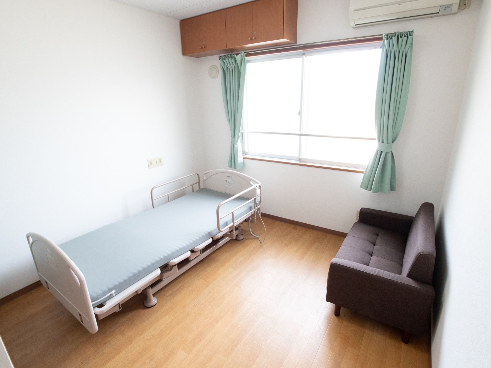 室内には、介護用のベッドとソファーが1台ずつ置かれている。カーテンがかかった窓の上には、エアコンと吊戸棚が設置してある。