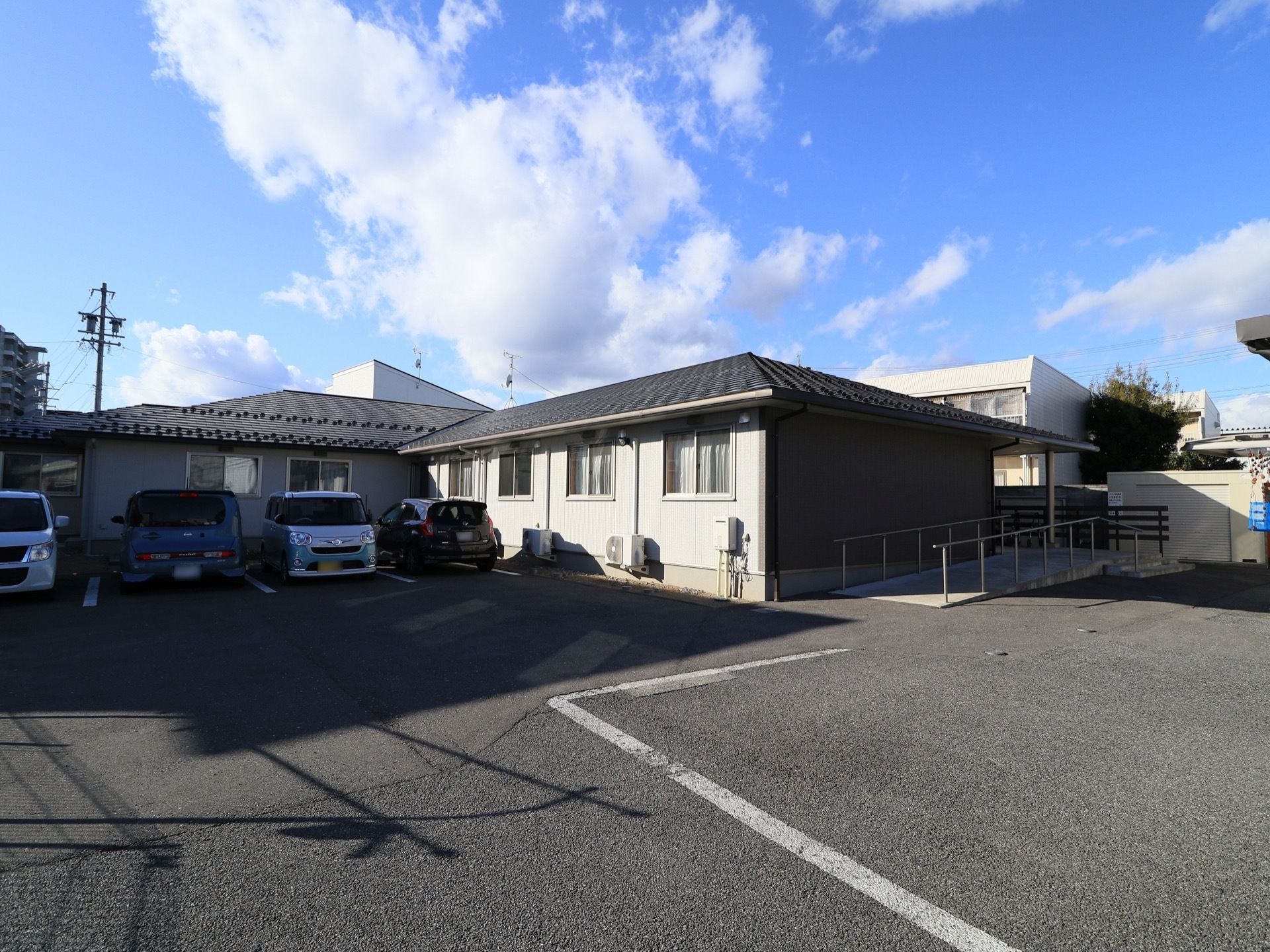 駐車場やスロープを完備している平屋建て施設の様子である。4台の車が停車している。青空が広がり清々しい。