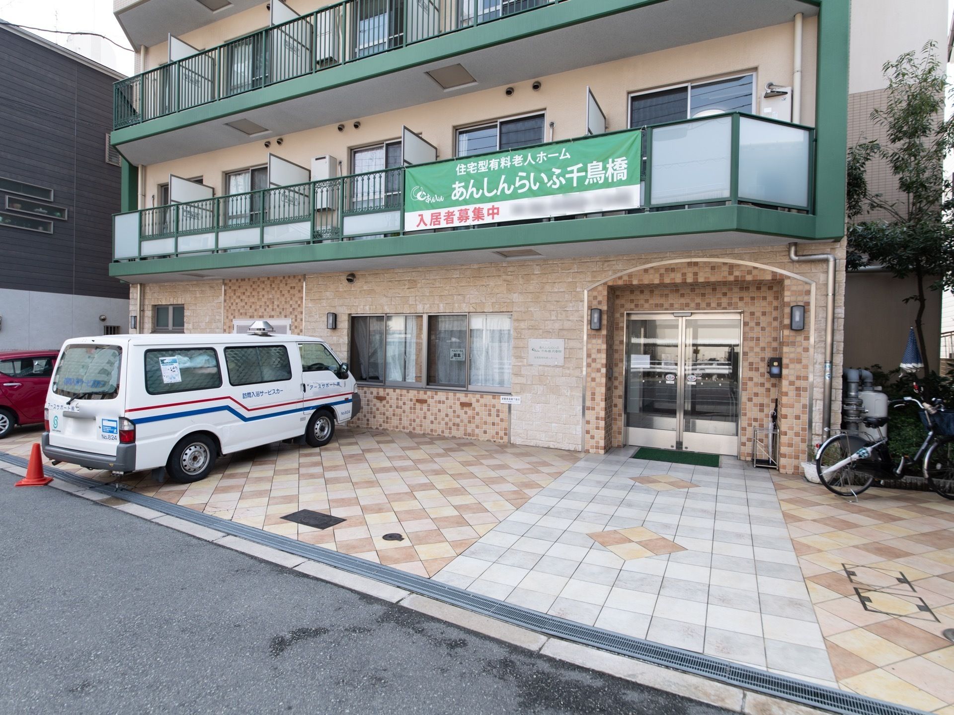 施設はマンションのような外観で、正面からは居室の窓とベランダがみえる。１階に自動ドアの入口と駐車場がある。