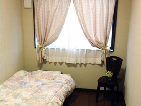 腰高の窓にピンク色のカーテンがかけられ、その手前に背もたれのある椅子が置かれている居室