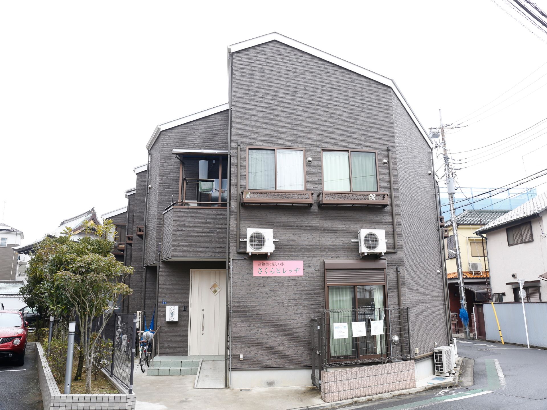 ダークグレーの外観にピンク色の看板掛けられている。木製の白い扉へはスロープが設置されている。自転車を停めることができる。