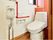 洋式便器が一据ある。温水洗浄便座が用意されている。赤色のＬ字型手すりが設けられている。その隣には小さな手洗い器が設置されている。