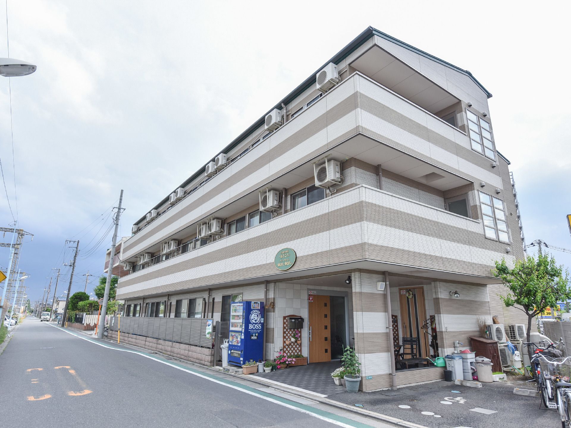 建物の外には自動販売機が設置されている。車や自転車を停めるスペースがある。植木鉢には花が植えられている。