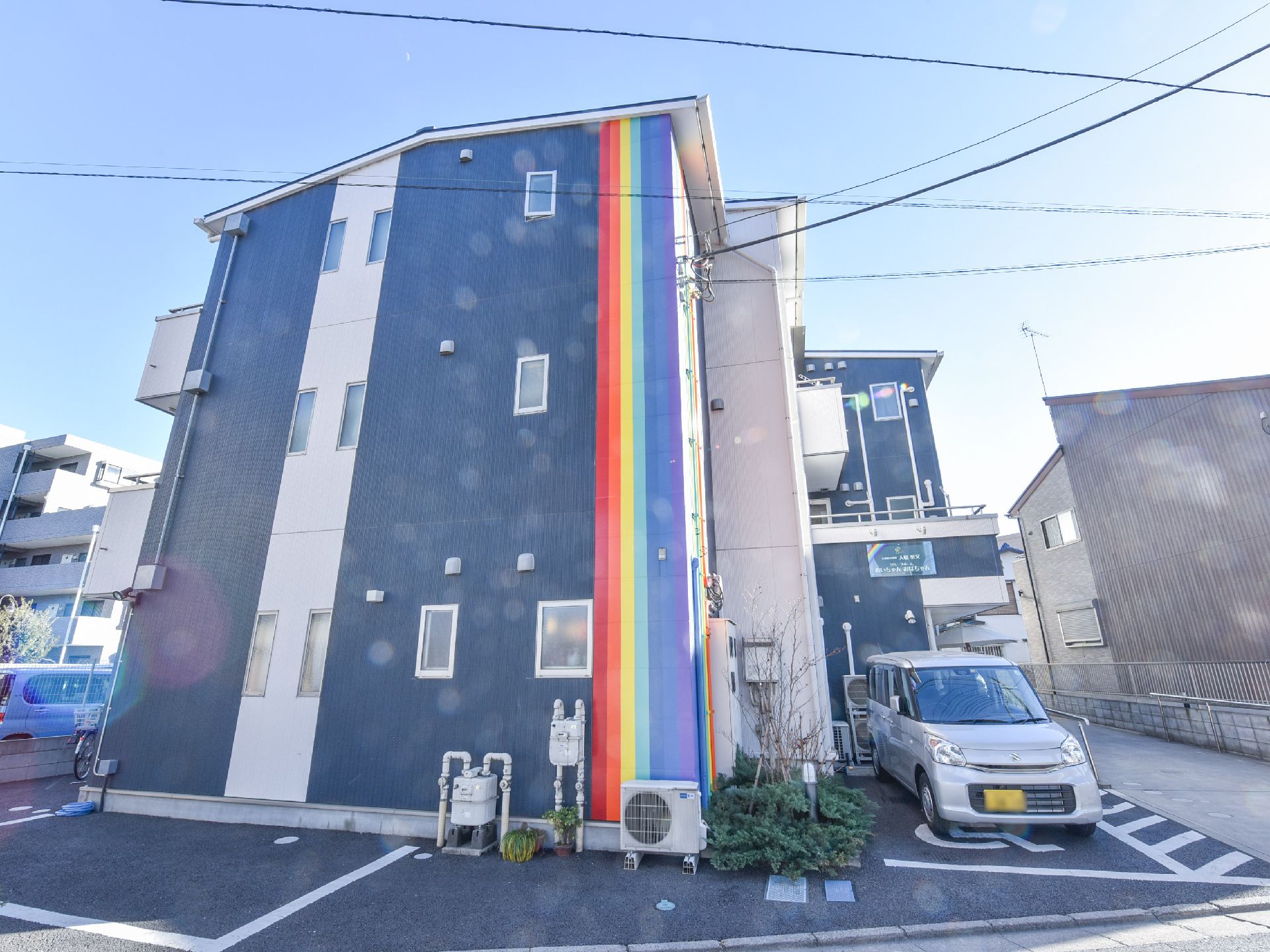 青色と白色で塗装された建物には、縦に7色の虹色のストライプ柄がはいっている。また、建物の脇には駐車場がある。