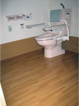 トイレの温水洗浄便座には背もたれ、手すりが設置され、壁には緊急呼出ボタンが設けられている。床はフローリングになっている。