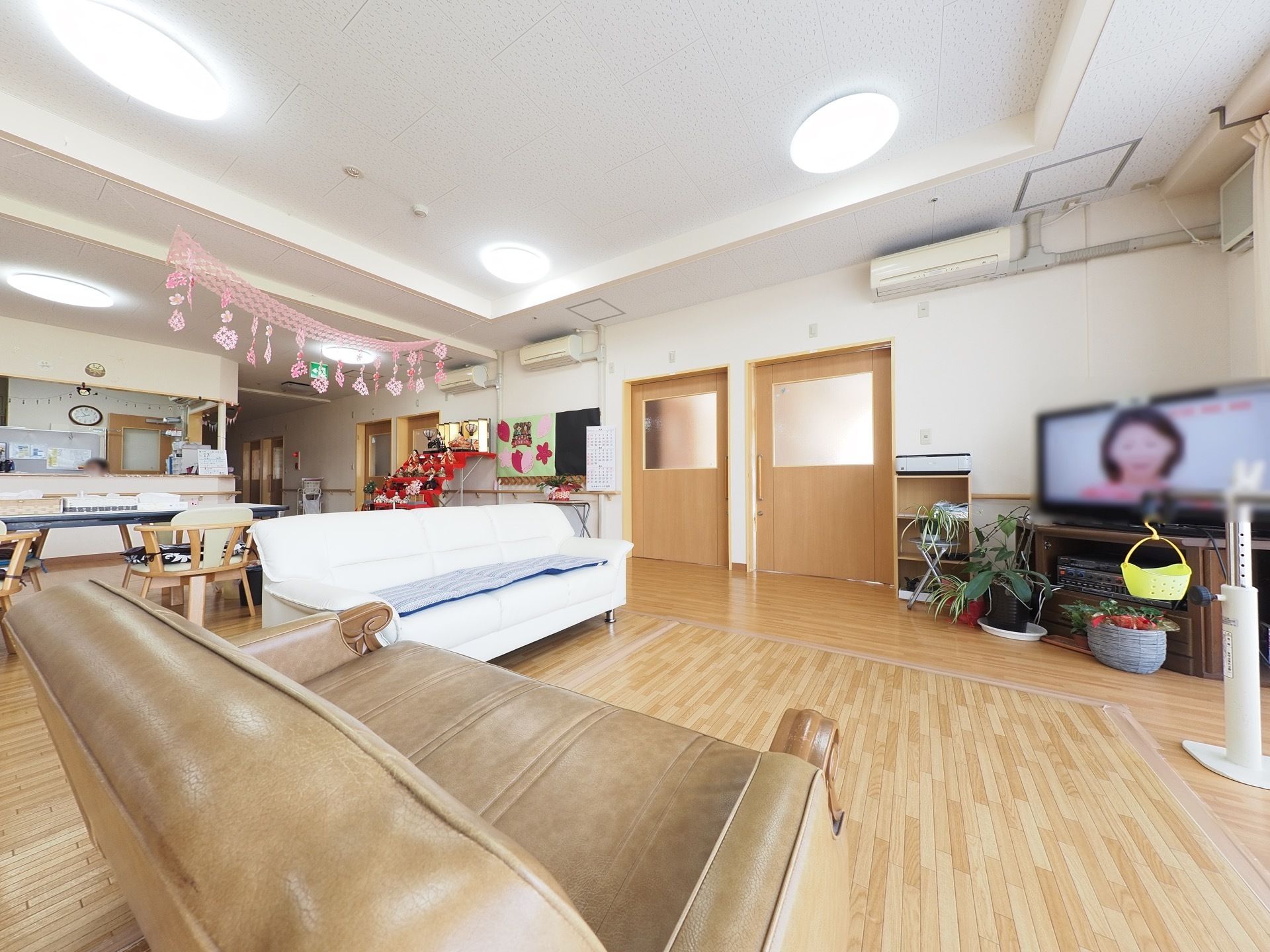 広い部屋に茶色と白色のソファが置かれており、壁際に大型テレビが設置されている。床はフローリングになっている。