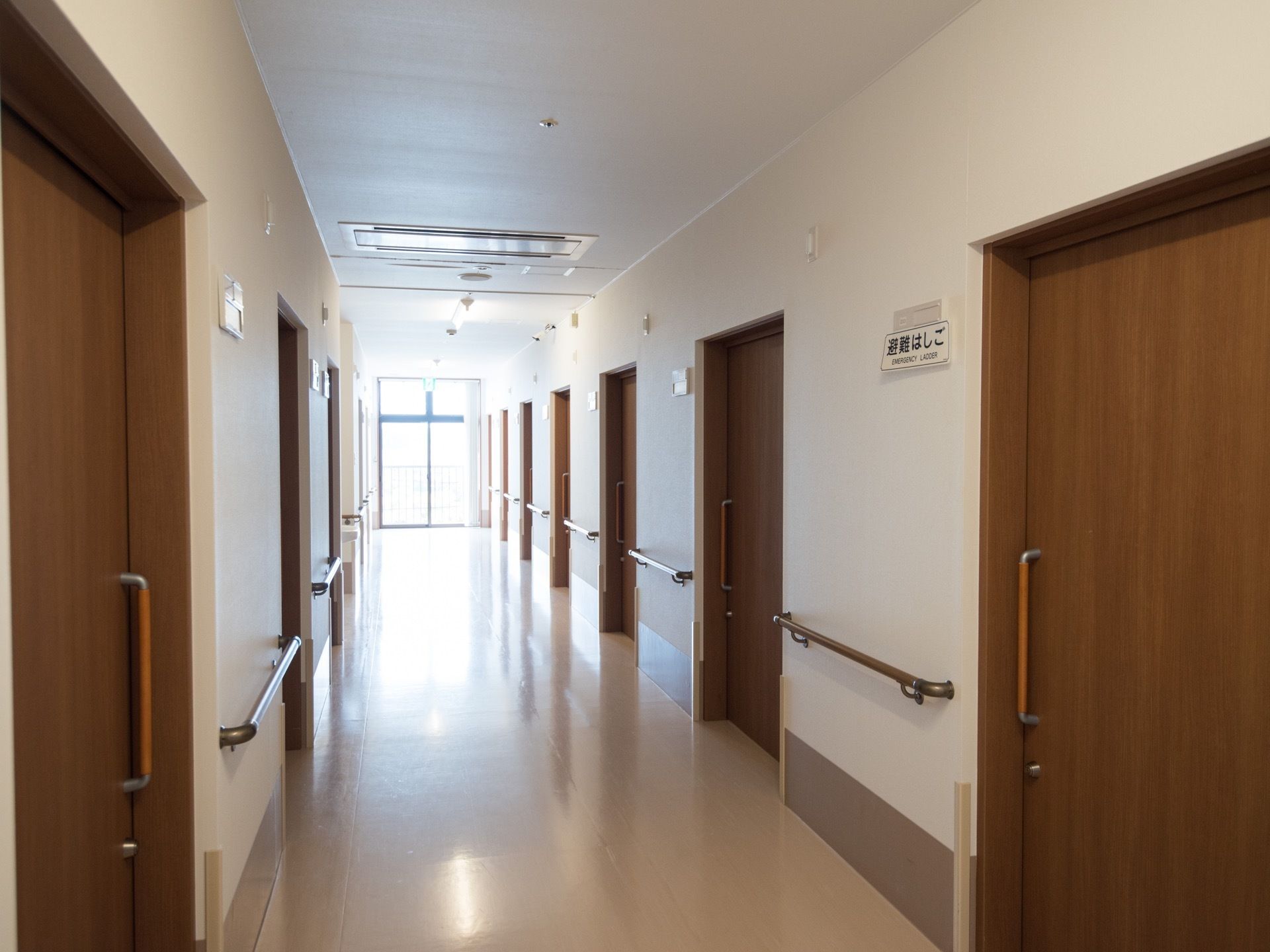 通路の両側の壁面に茶色い引き戸が並ぶ、落ち着いた雰囲気の廊下である。突き当たりの非常口から外光が射し込んでいる。