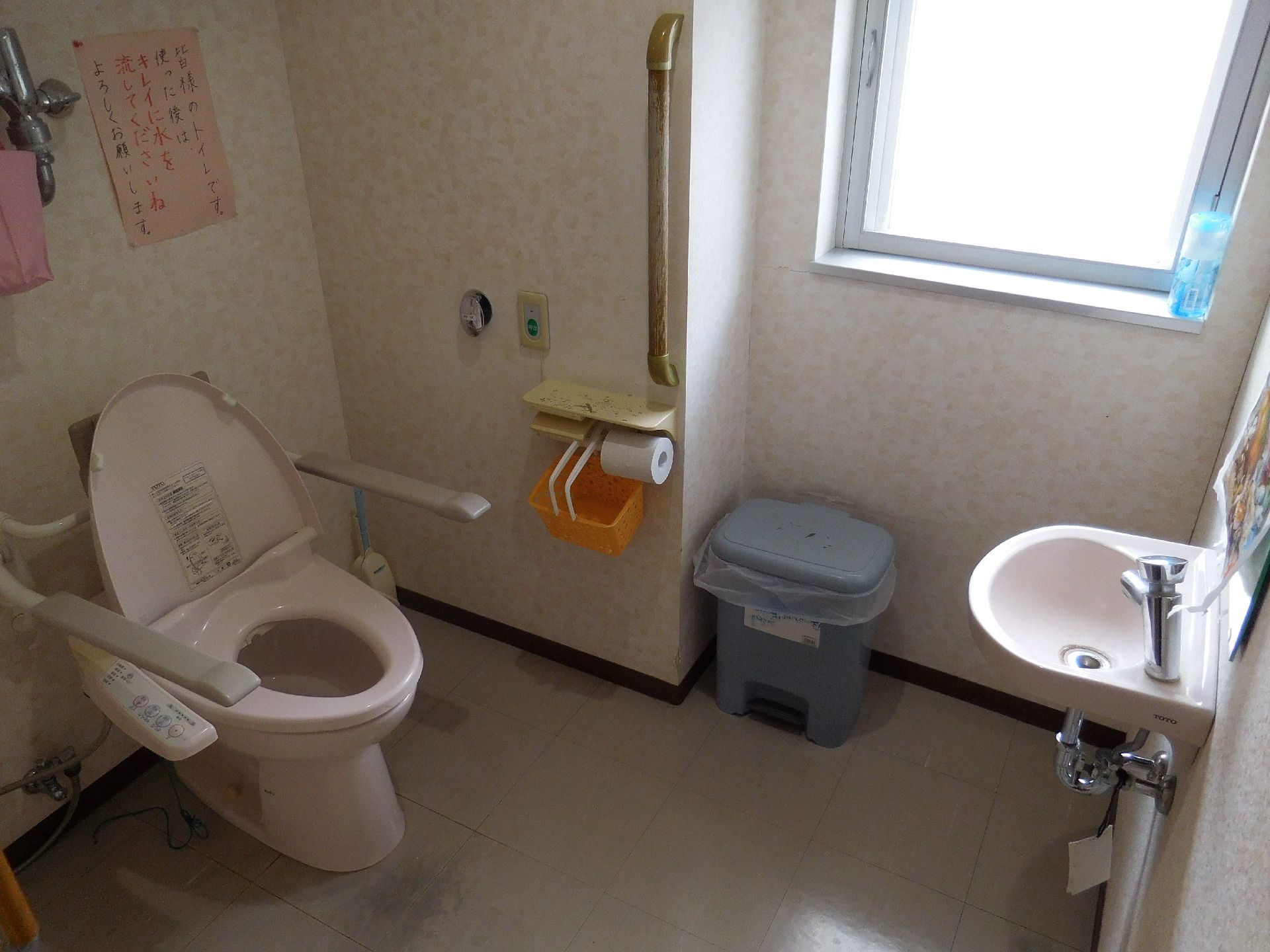 施設のトイレはウォシュレットの水洗トイレ。便器には可動式の手すりが外付けされている。壁にも手すりがあり、小型の洗面台も備え付けられている。