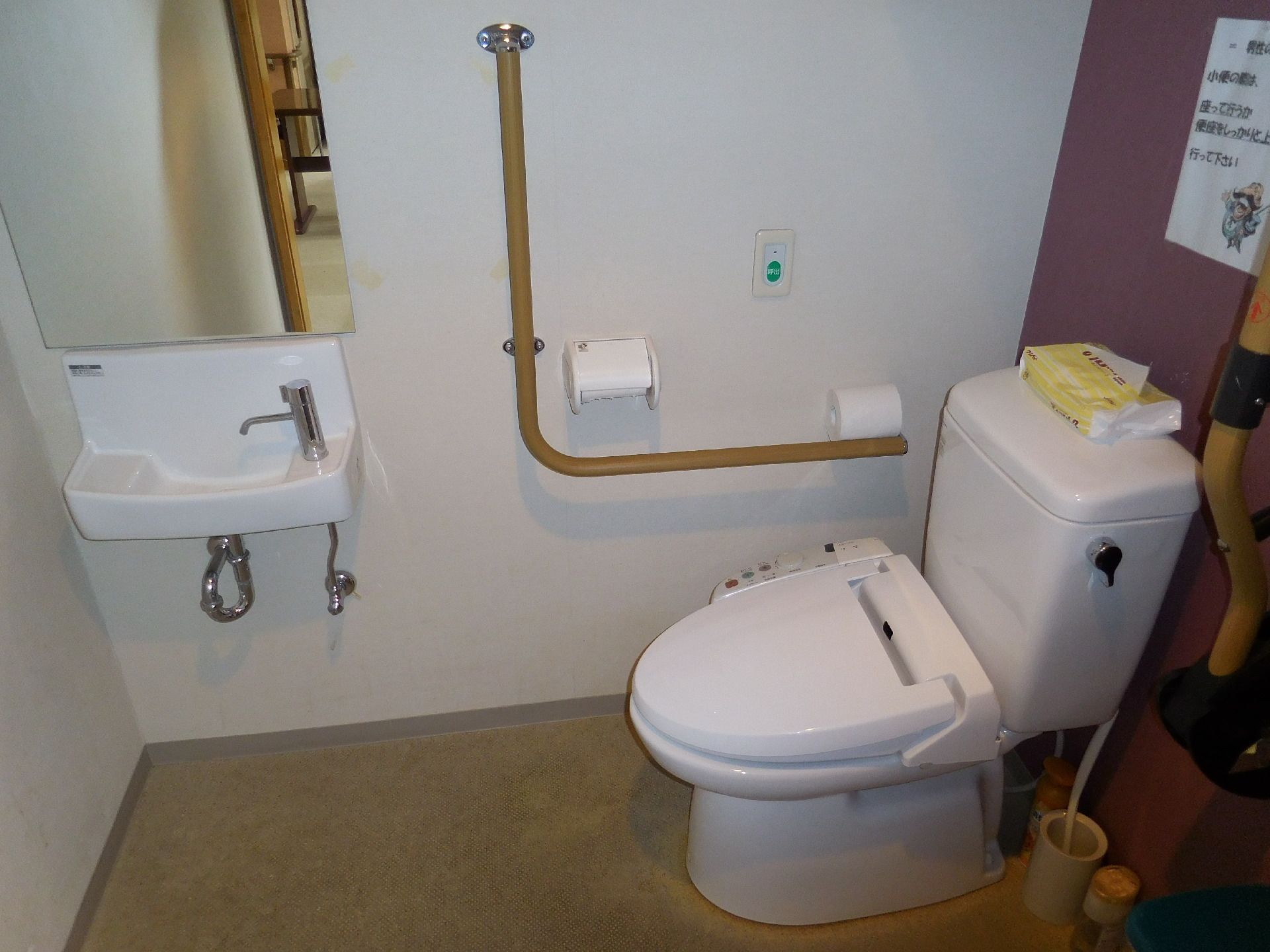 水洗トイレの横には可動式の手すりも設置されている。壁には貼り紙があり、予備のペーパーなどの備品も置かれている。