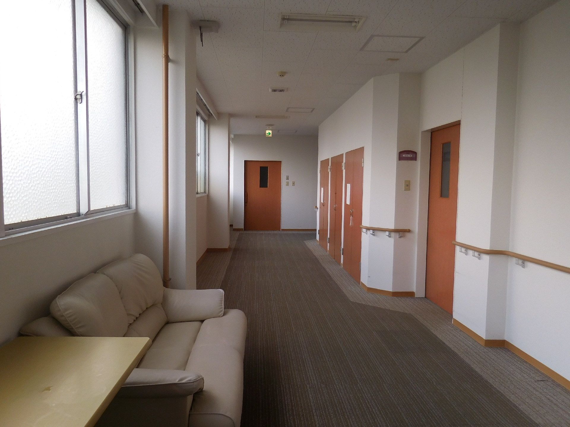 施設の廊下は白壁にグレーの絨毯。幅広で茶色の扉が並んでいる。窓際にはソファーやテーブルなども置かれている。