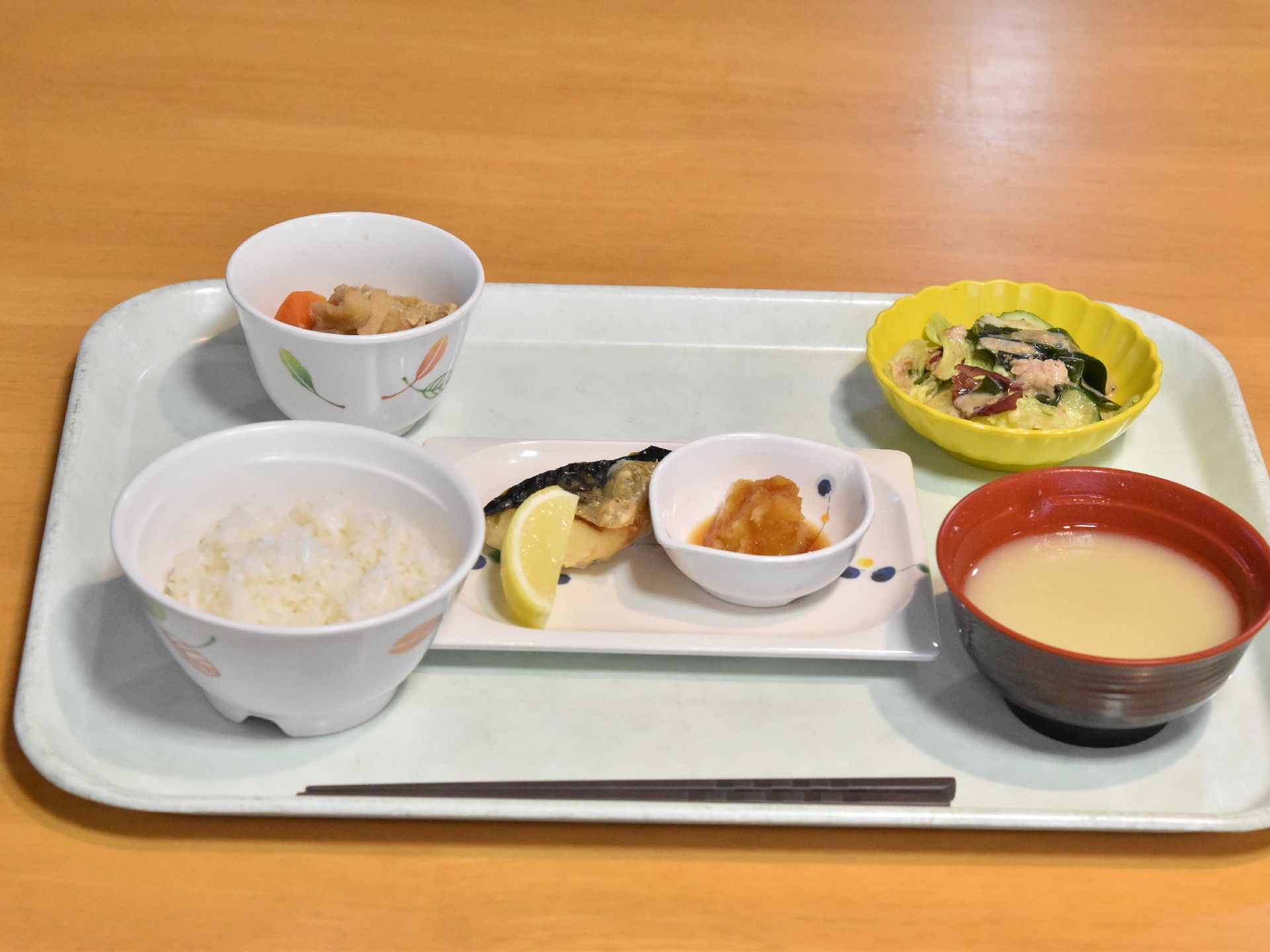 茶色のテーブルに水色のトレイで食事が配膳されている。箸があり、ご飯と味噌汁、サラダと肉じゃが、焼き魚にレモン、大根おろしが乗せられている。