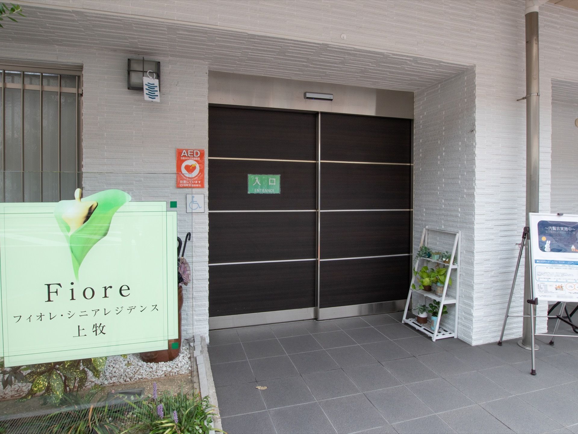 茶色い大きな扉の自動ドアの横に植物が飾られた白いラックが置いてある。植栽スペースに施設名が書かれた緑色の看板が設置されている。