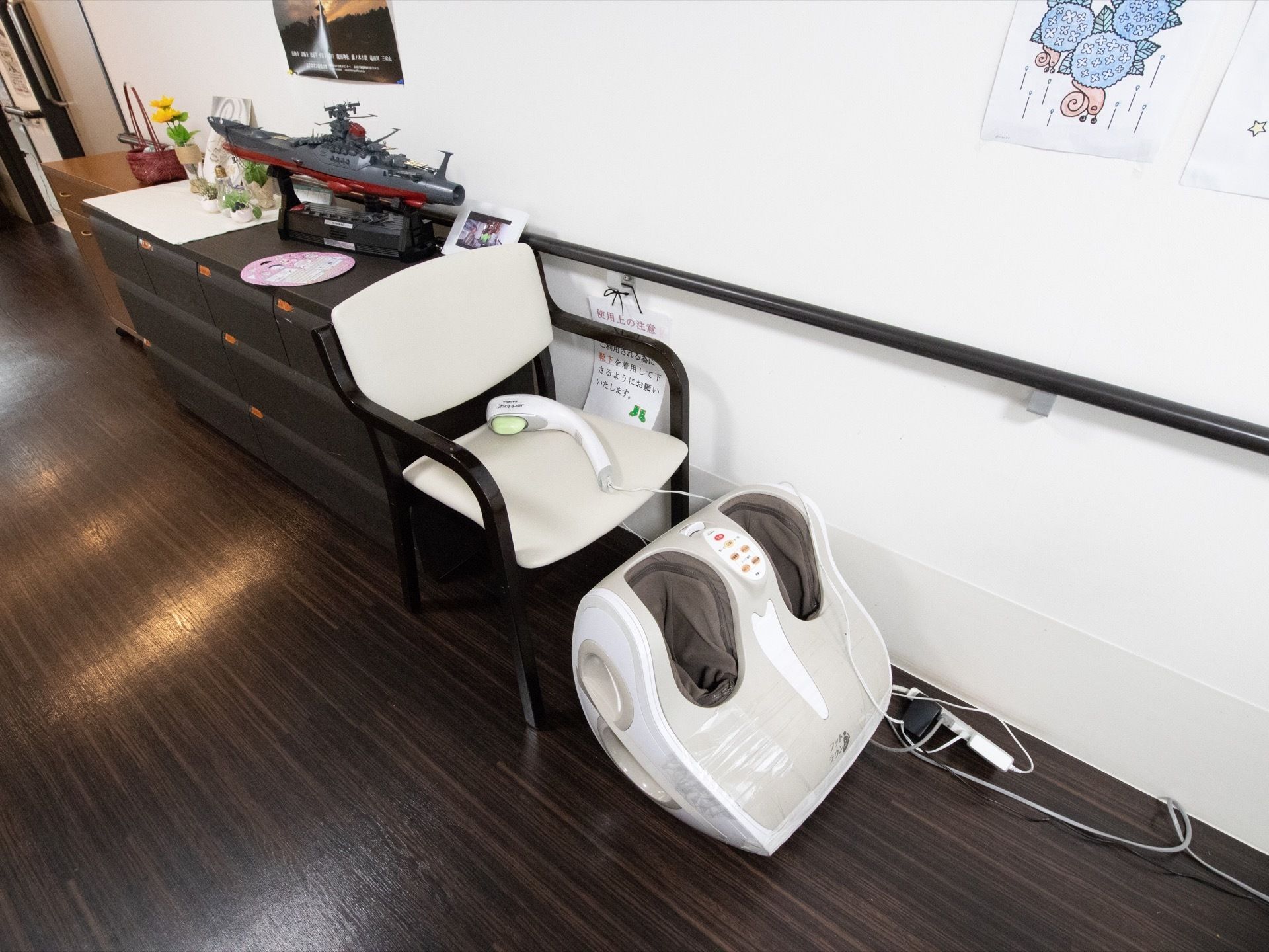 軍艦のプラモデルが飾られた茶色いチェストが置いてある。チェストの横に椅子があり、足元にマッサージ器が置いてある。