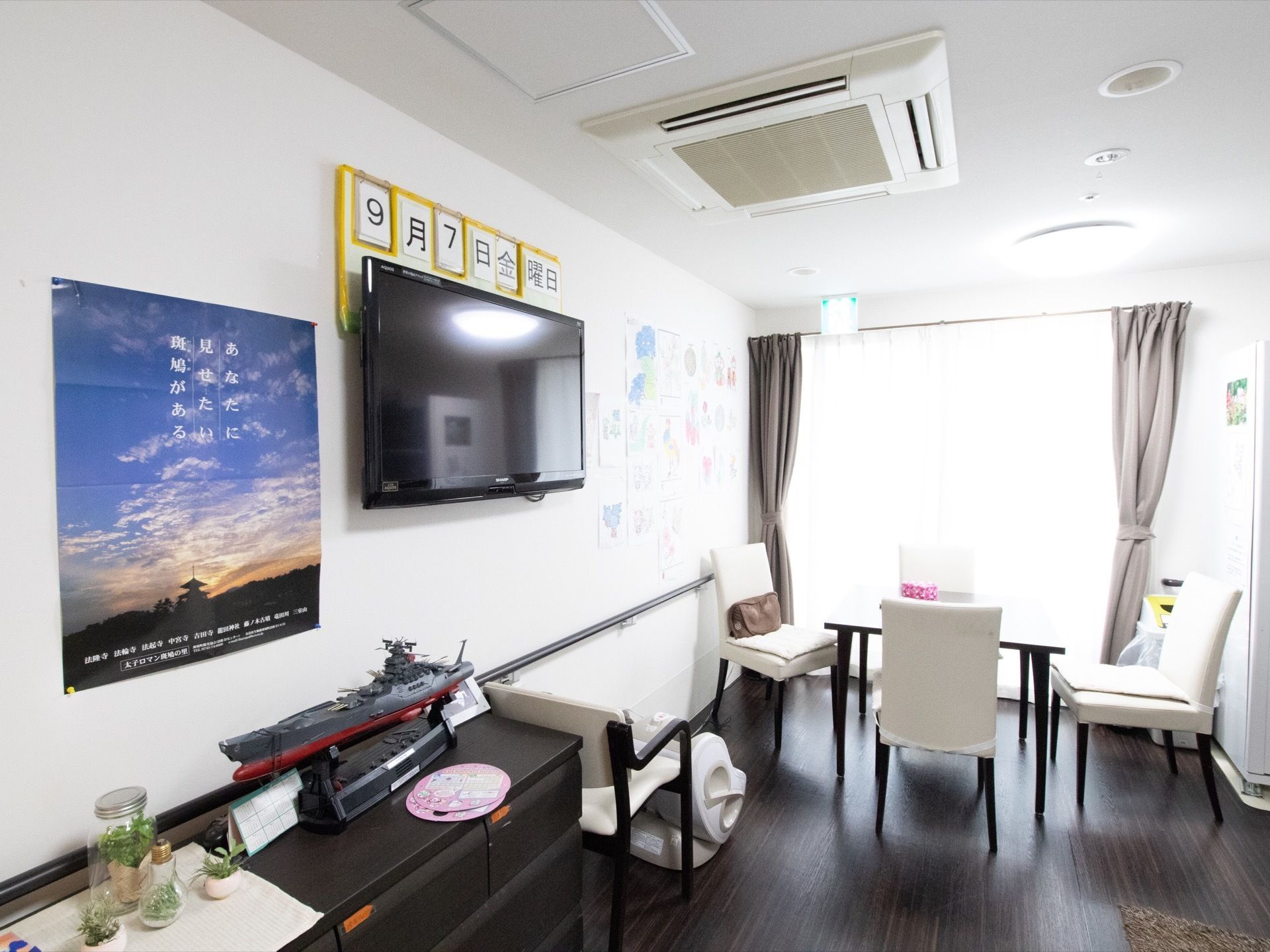 壁に大きな黒い薄型テレビが掛けられている。テレビの上には大きな文字で日付が表示されている。１卓の机と４脚の椅子が置いてある。