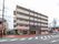 ５階建てのマンションのような白い建物が建っている。施設前の歩道はオレンジ色に塗られ、車道を１台の軽自動車が走っている。