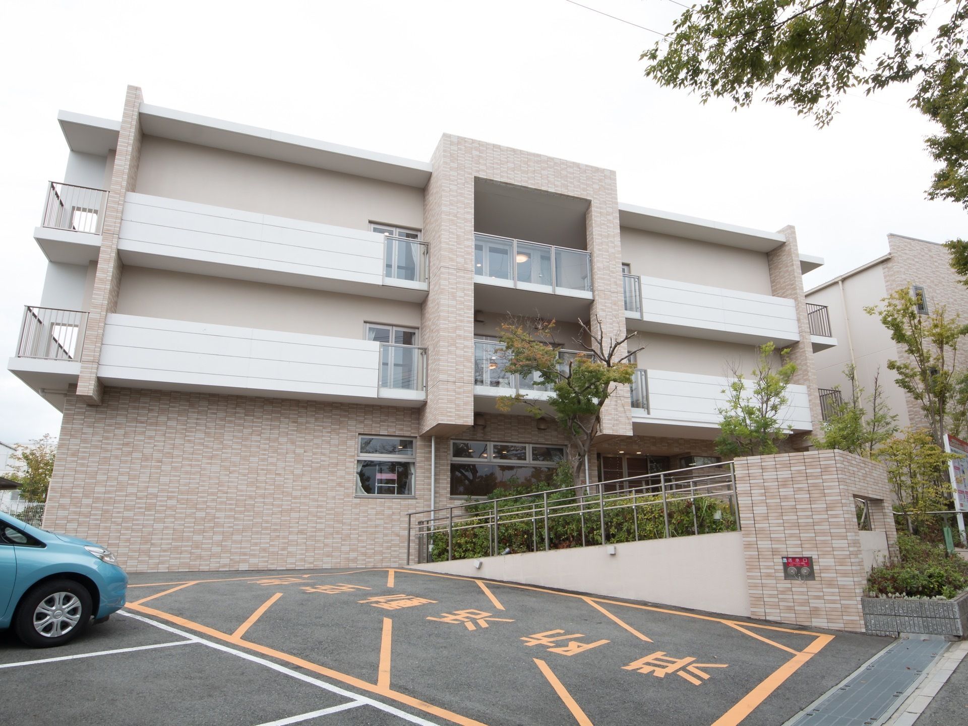 淡いレンガ色と白の外壁が落ち着いた佇まいの、鉄筋コンクリート造り３階建ての建物。入口付近の駐車場から玄関まで緩やかなスロープになっている。