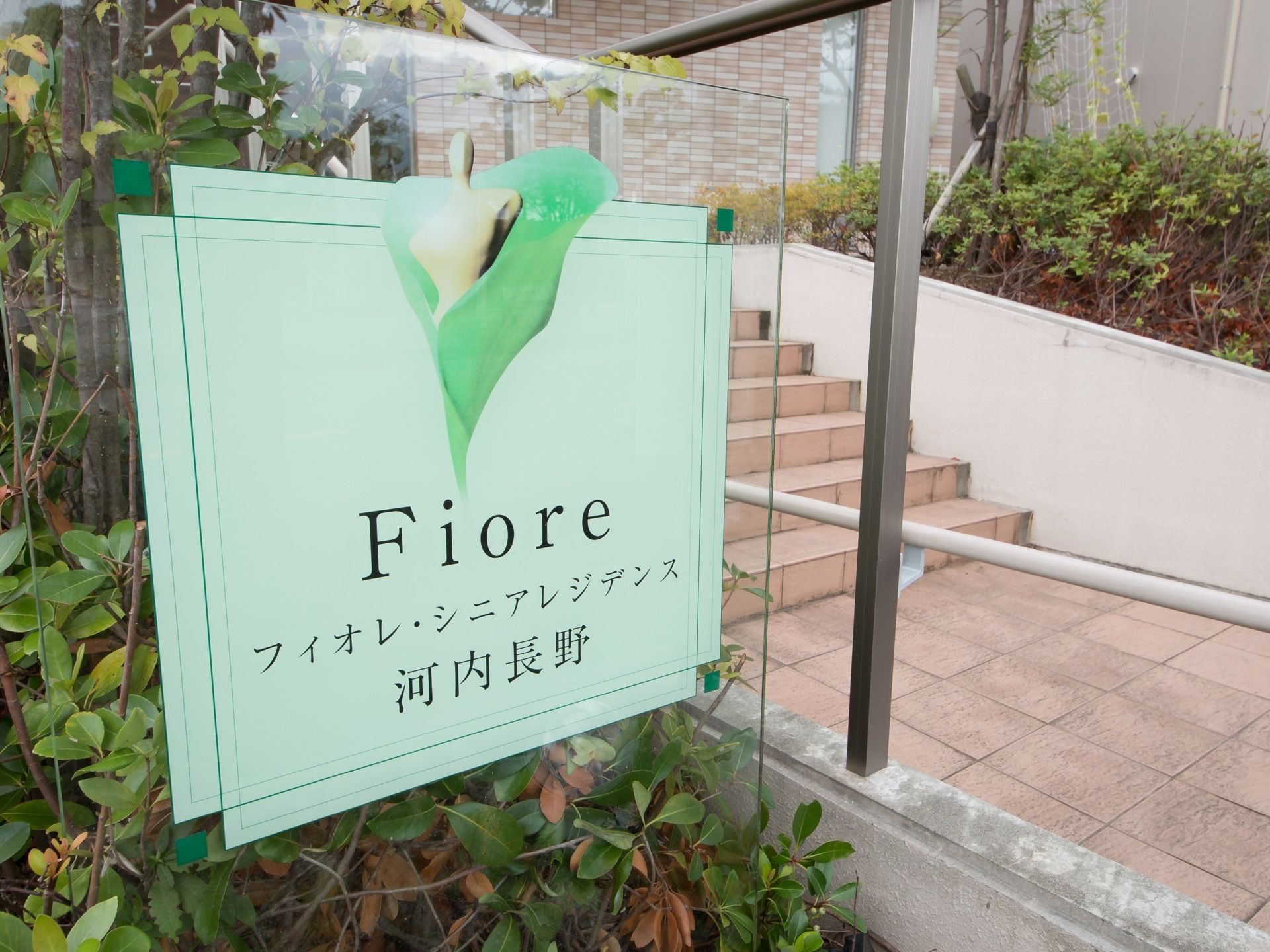 玄関前まで続く通路にはレンガが敷かれており、両側に植え込みが配置されている。植物の間に置かれた透明なパネルに施設名とロゴが書かれている。