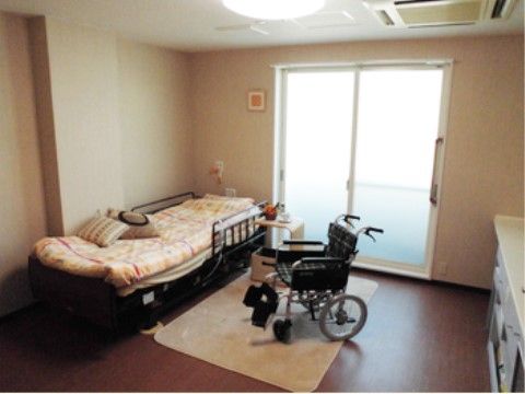 バリアフリーの茶色い床に白いマットが敷かれ、その上に車椅子が置いてある。ベッドには柵が設置され、横にはサイドテーブルがある。