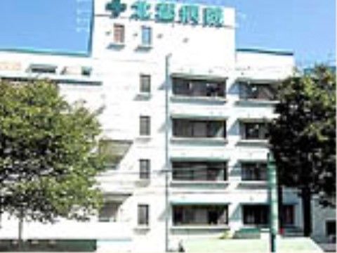白い建物の病院がある。建物の一番高いところに、緑色で病院名が書かれている。病院の前には木が植えられている。