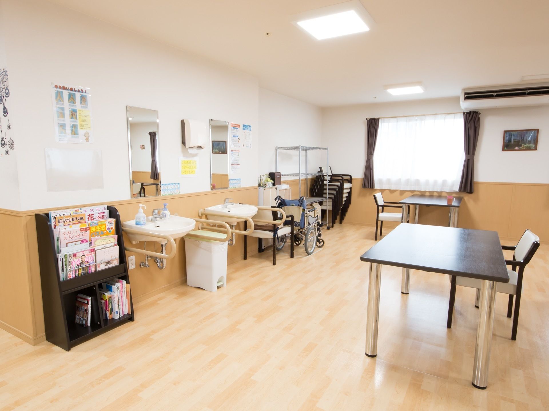 明るい色調の談話室。大きなテーブルと椅子が並んでいる。壁際には洗面台が２台設置され、マガジンラックには雑誌や本が置かれている。