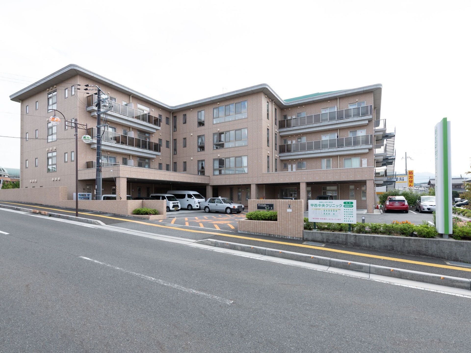 広い道路に面した敷地に、４階建ての建物が建っている。駐車場があり、タクシーや自家用車が停まっている。