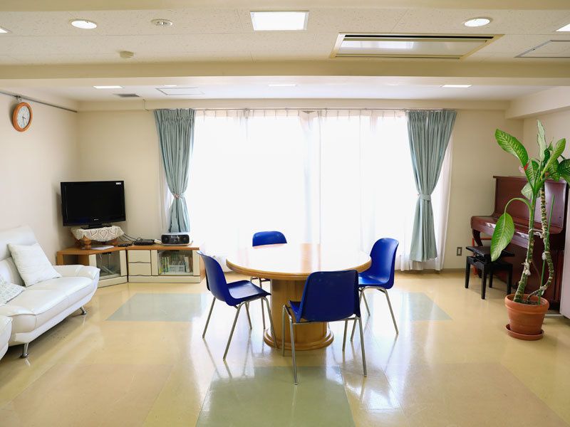 白を基調とした空間。壁際にはピアノが置かれている。中央には木製の丸テーブルと青い椅子が配置されている。