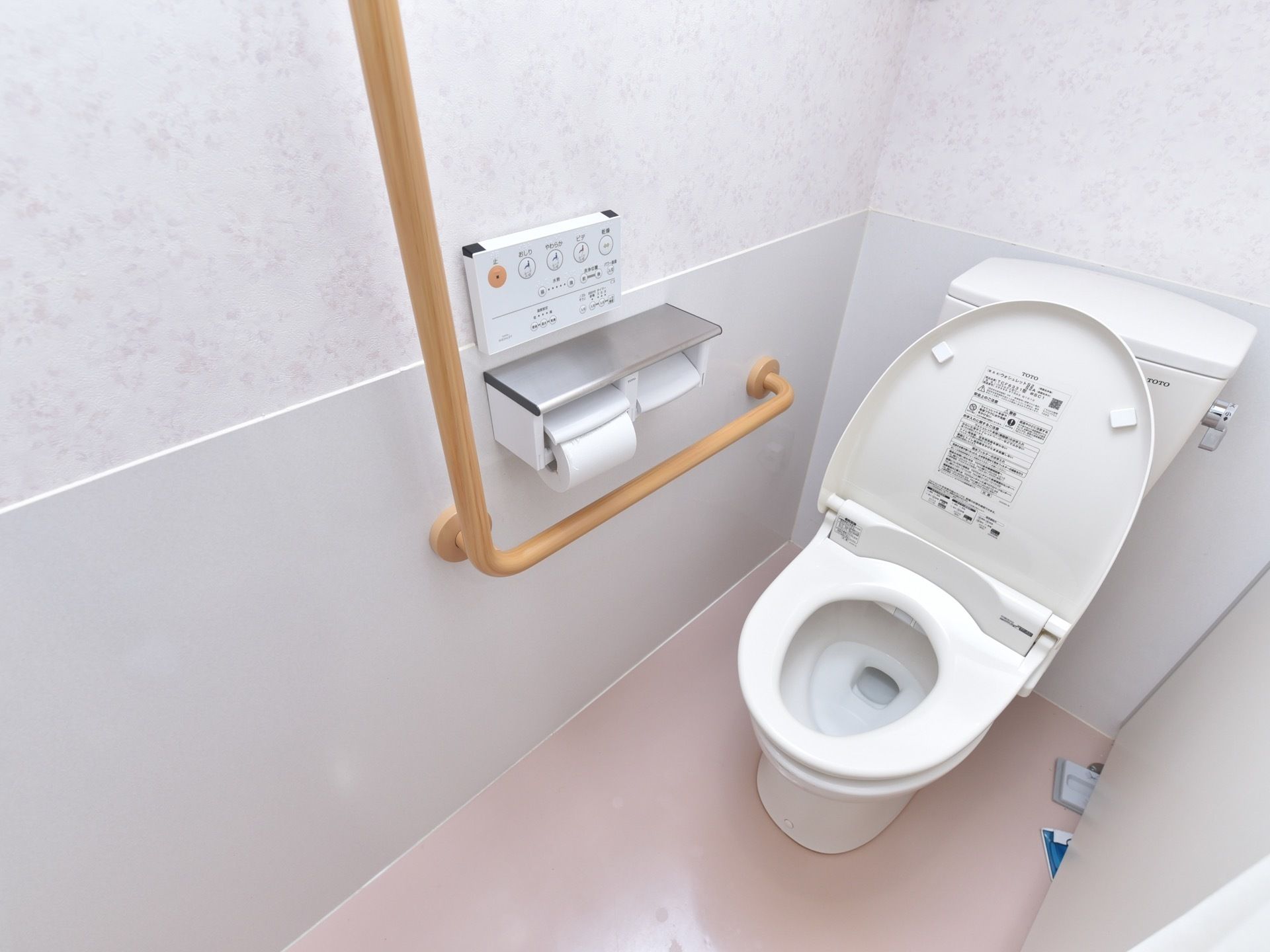 全体が白でまとめられたトイレで、洗浄機能付き便座、L字型手すり、トイレットペーパーホルダーが設置されている。
