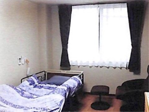 腰高窓から明るい光が差している。窓には白いレースカーテンとこげ茶色の遮光カーテンが掛かっている。介護ベッドが置いてある。