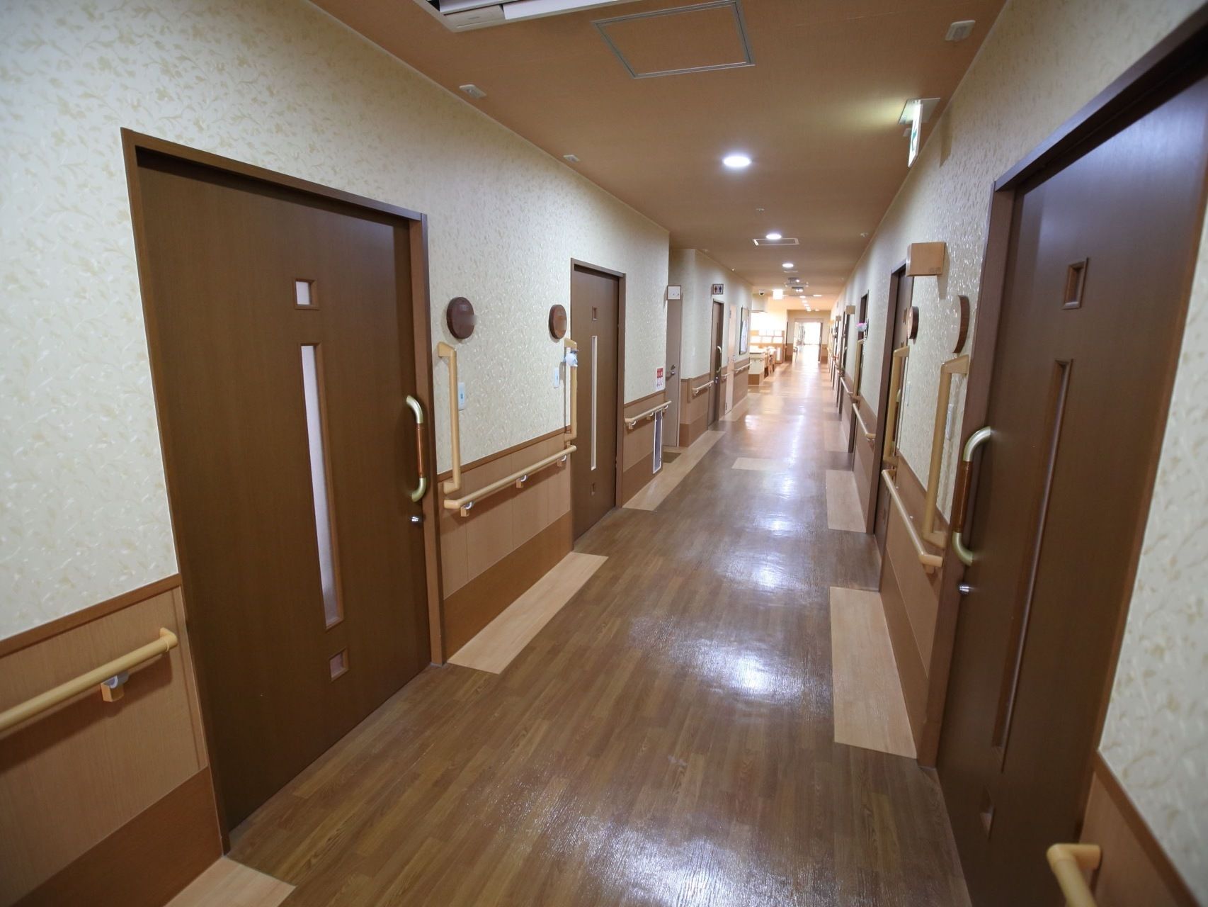 ブラウン系で統一された居室前の廊下である。直線で見通しがよく障害物は置かれていない。両サイドの壁には手すりが設置されている。