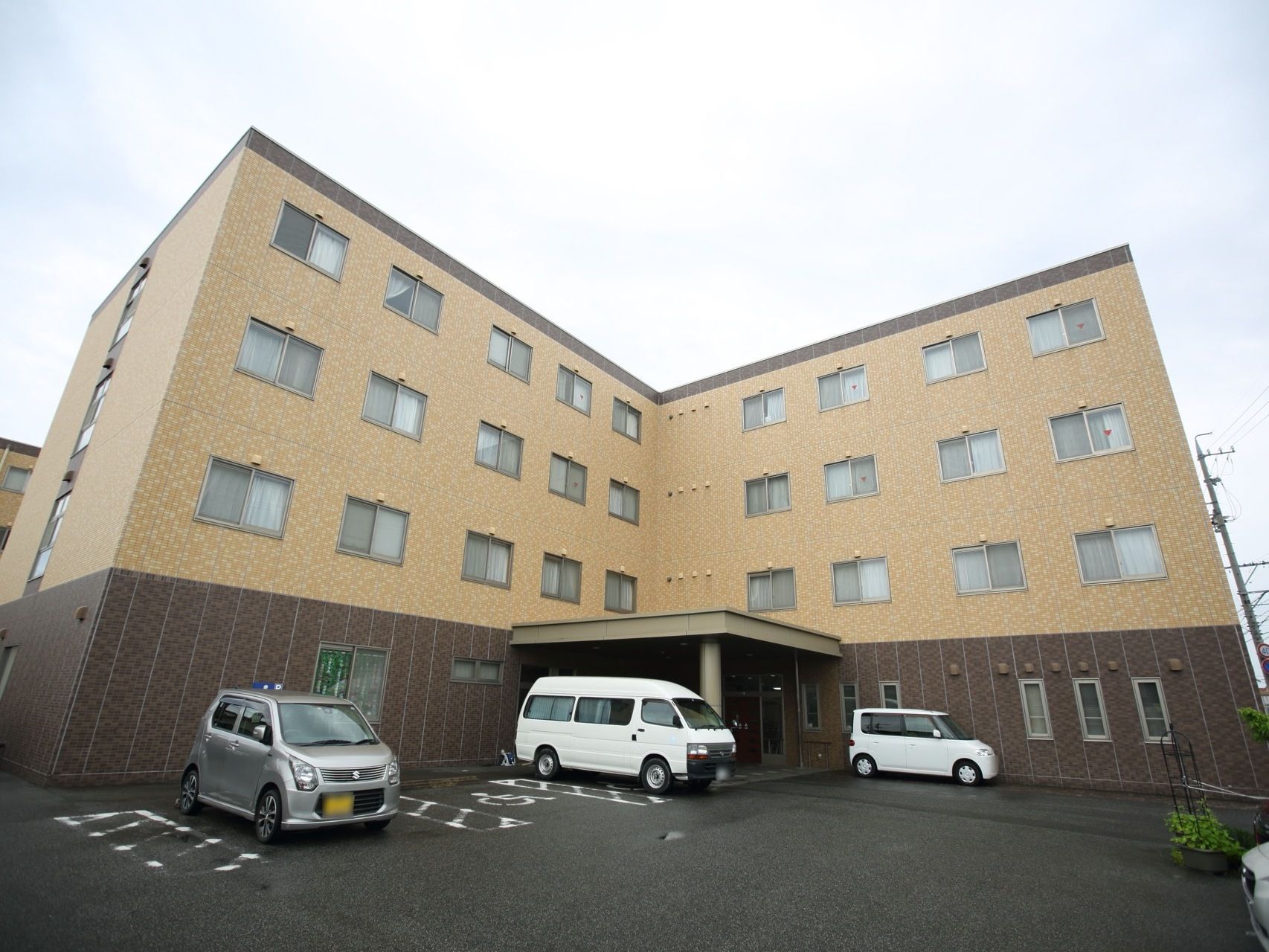 ベージュとブラウンのツートーンの外壁が印象的な４階建て施設の外観である。駐車場を完備し、乗用車が3台停車している。