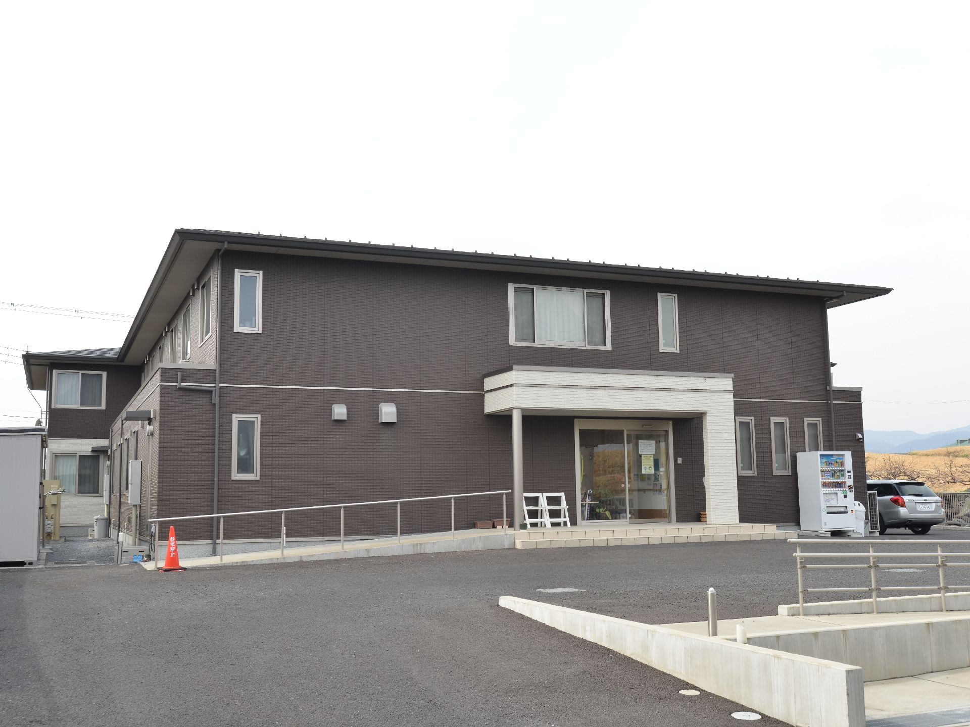2階建てのシンプルな雰囲気の建物。周りは自然に囲まれた好立地。建物周辺には自動販売機と駐車場がある。
