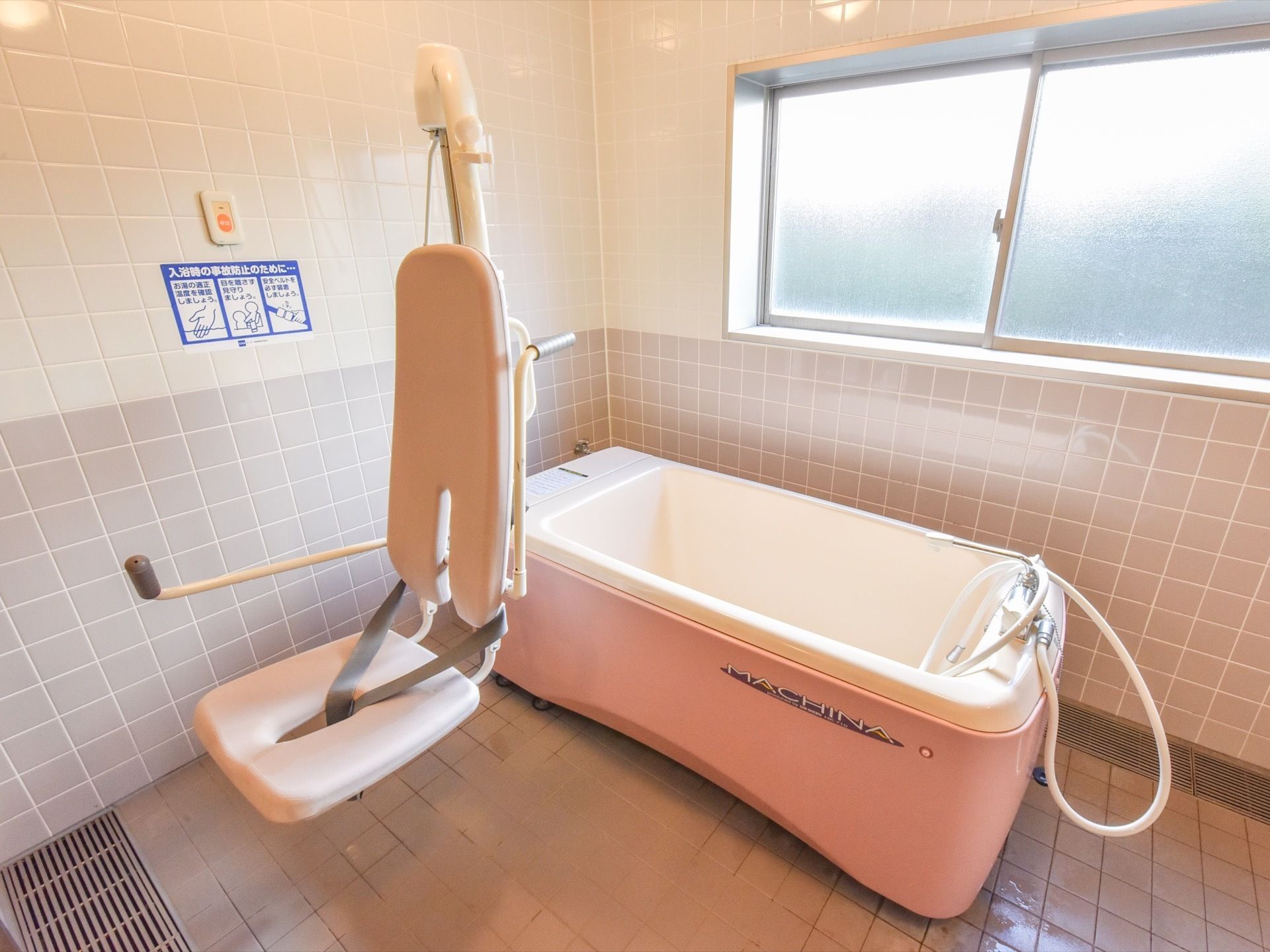 ベージュ色のタイル張りの浴室には、介護用の浴槽が備えられている。座ったまま入浴が可能である。事故防止のための案内文書が貼られている。