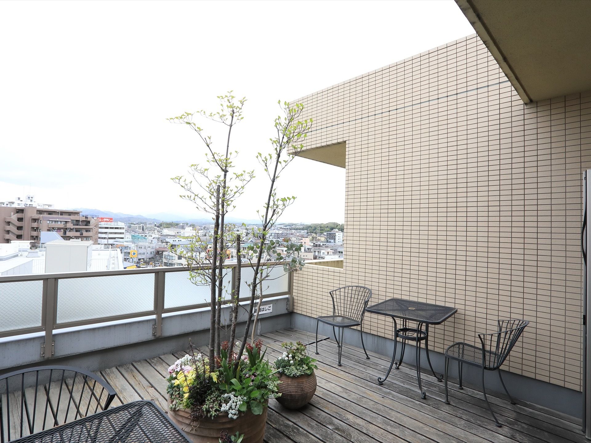 見晴らしの良いウッドデッキ風テラス。テーブルセットが配置されている。花や木が植えられている鉢が置いてある。