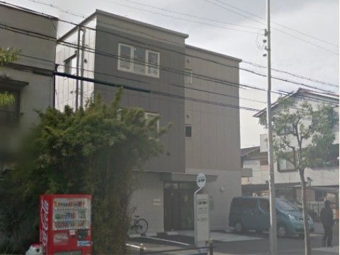 建物の前は公道にっており、すぐ近くにバス停がある。隣接する住宅前に植え込みがあり、その前に自動販売機が設置されている。