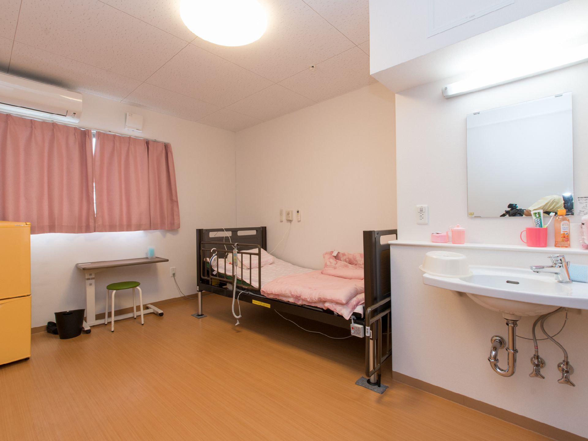白色の天井と壁。床はぬくもり感のある茶系の木目調。壁際にベッドが置いてあり、その隣には洗面台。窓があり、上部にはエアコンが設置されている。