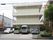 薄いクリーム色の3階建ての外観。建物の傍には5台の車が止められている。敷地入口の傍には庭木や緑が植えられている。