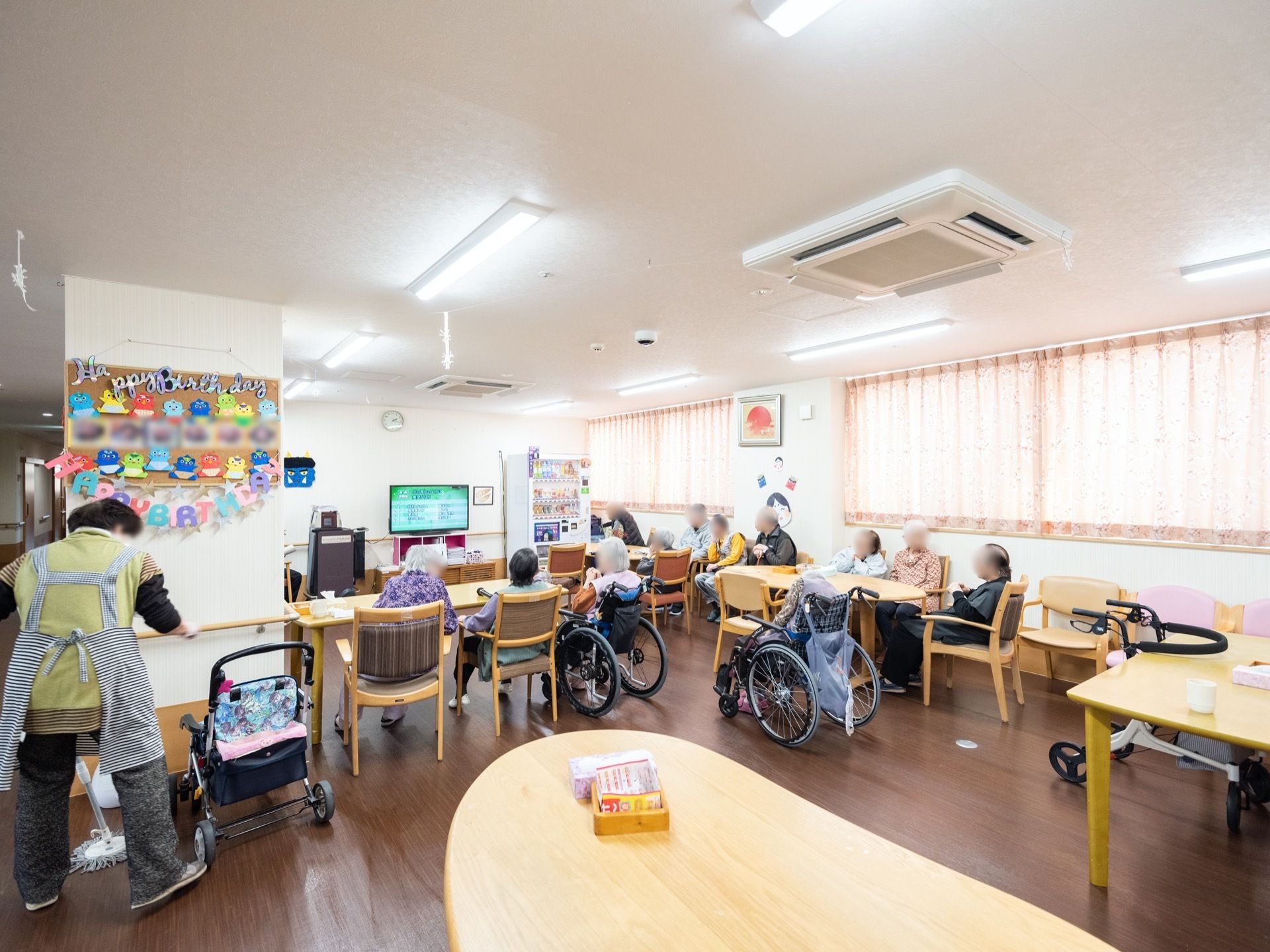 カラオケのある共有スペースである。入居者様たちがモニター画面を眺めている。脇では、スタッフが清掃作業中である。