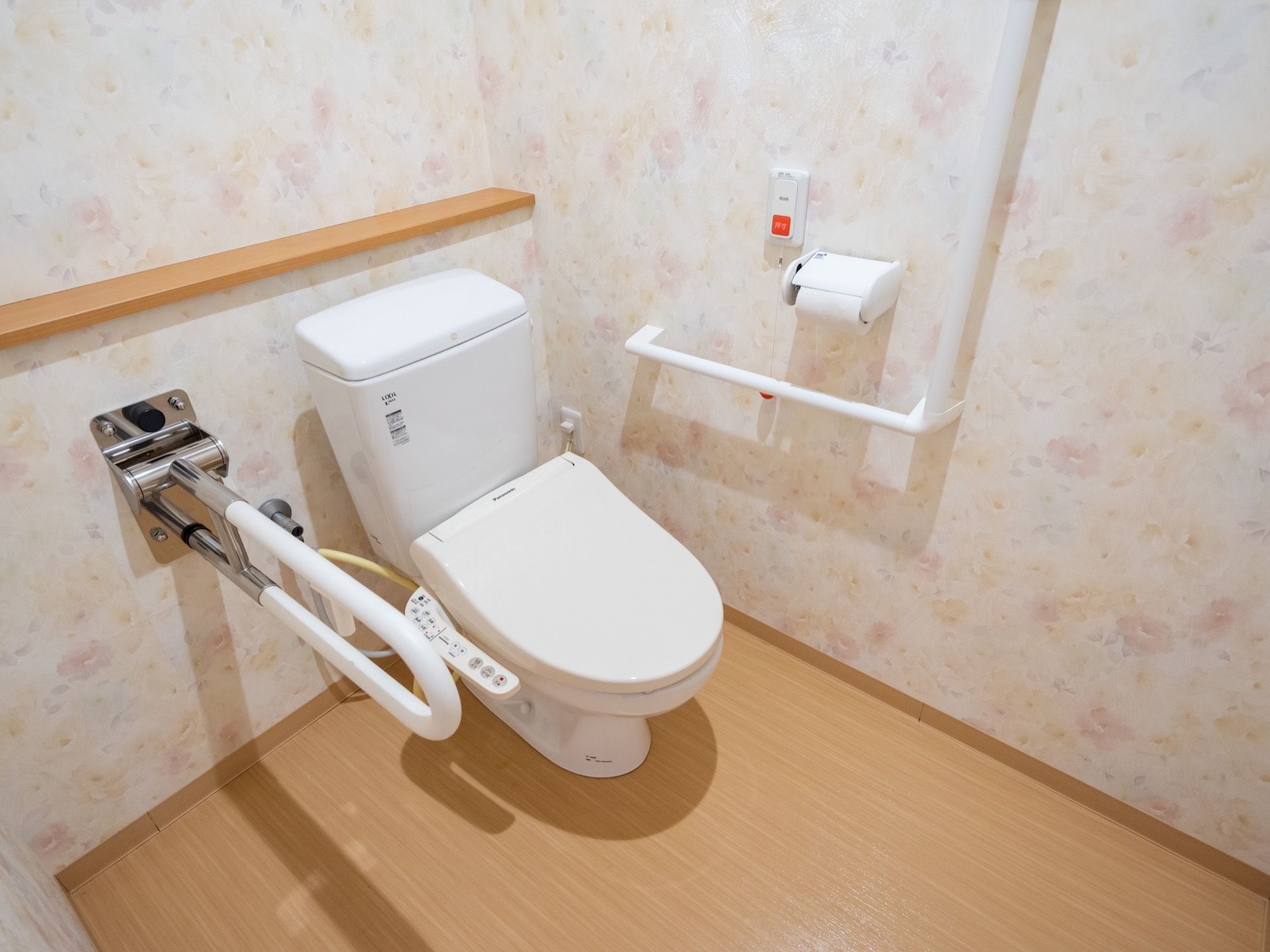 薄い花柄模様の壁紙が貼られたトイレである。洋式便器が設置され、両脇に手すりが付いている。壁にはナースコールなどがある。