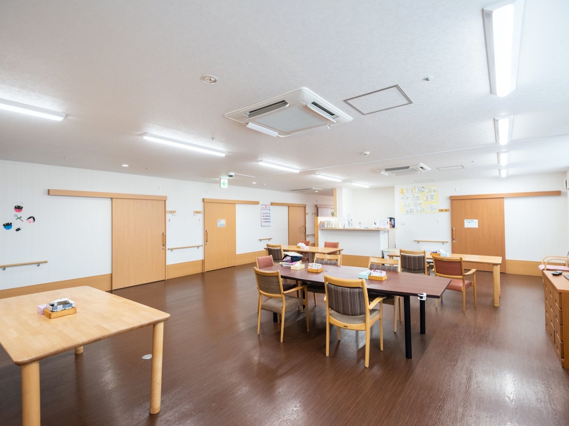 広いスペースの食堂である。木製のテーブルと椅子が並べられている。テーブルの間隔は広い。奥に厨房カウンターがある。