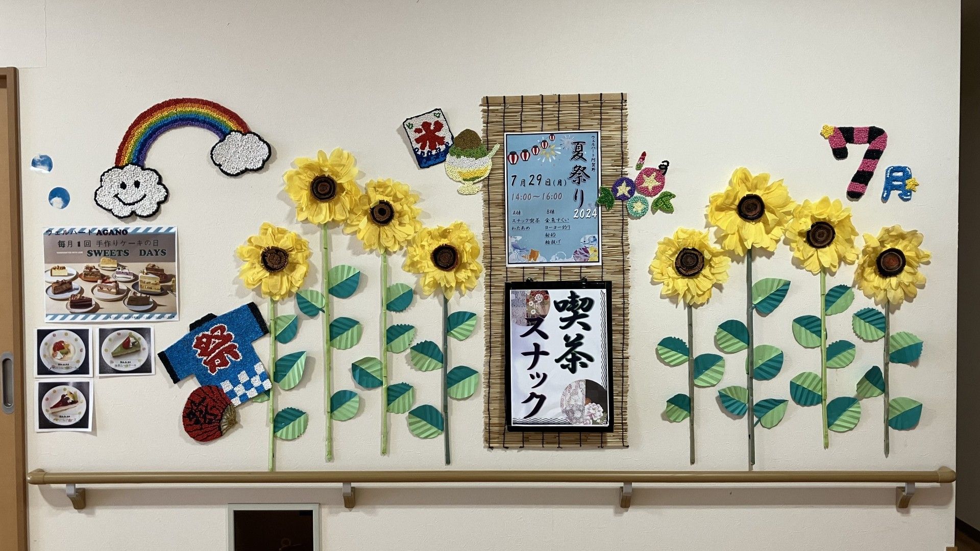 居室入口の横の壁に、入居者様の塗り絵作品が９枚貼り付けられている。居室の表札の下には折り紙で作られた花が飾られている。