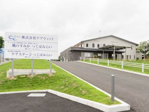 敷地内には施設の看板が立てかけられている。植え込みには緑が広がっている。施設は2階建てで、玄関入口には大きな屋根が設置されている。