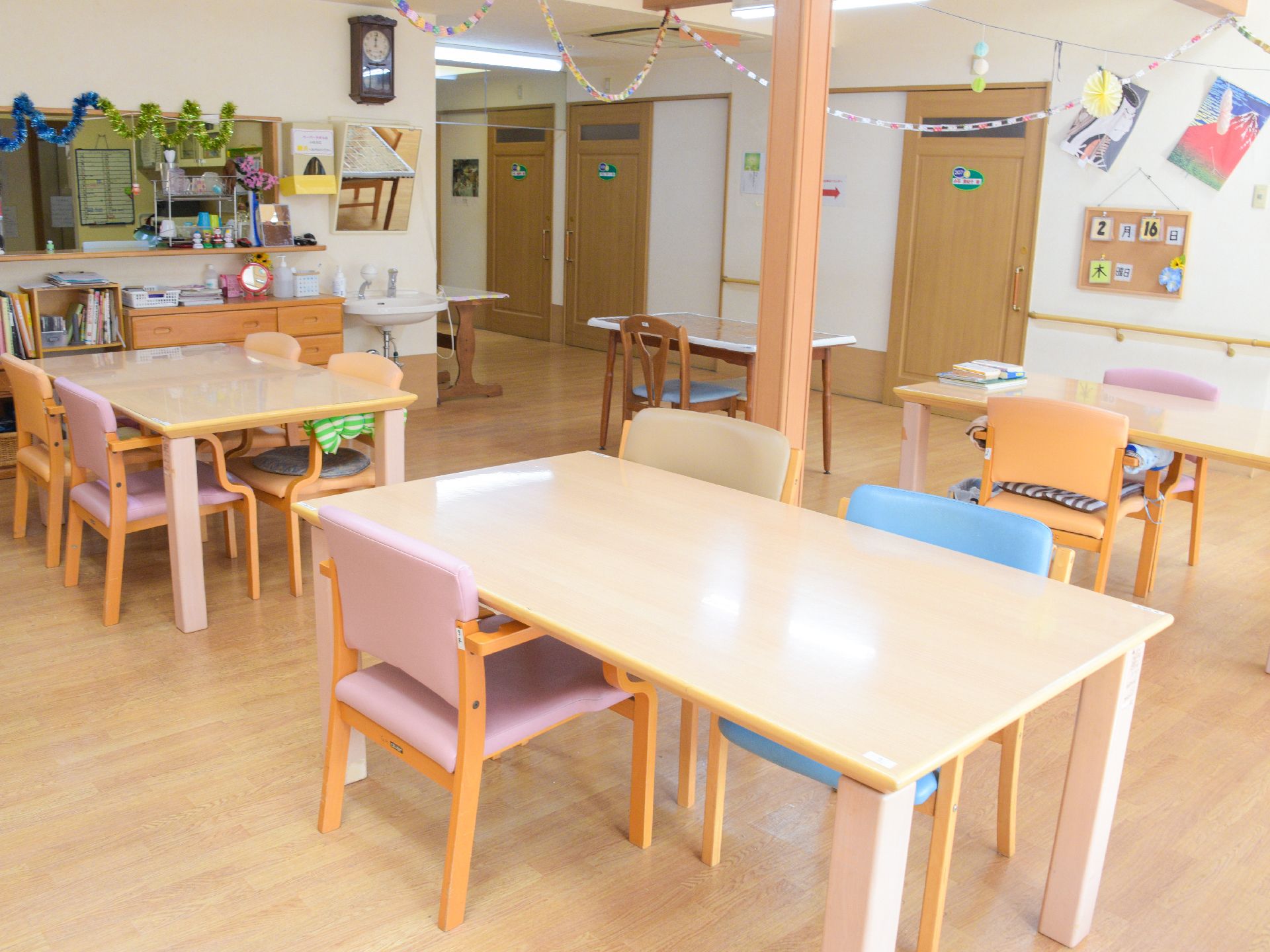 食堂には、本棚、洗面台、テーブル、椅子などが設置されている。天井や壁には、入居者様の手作りの品が飾ってある。